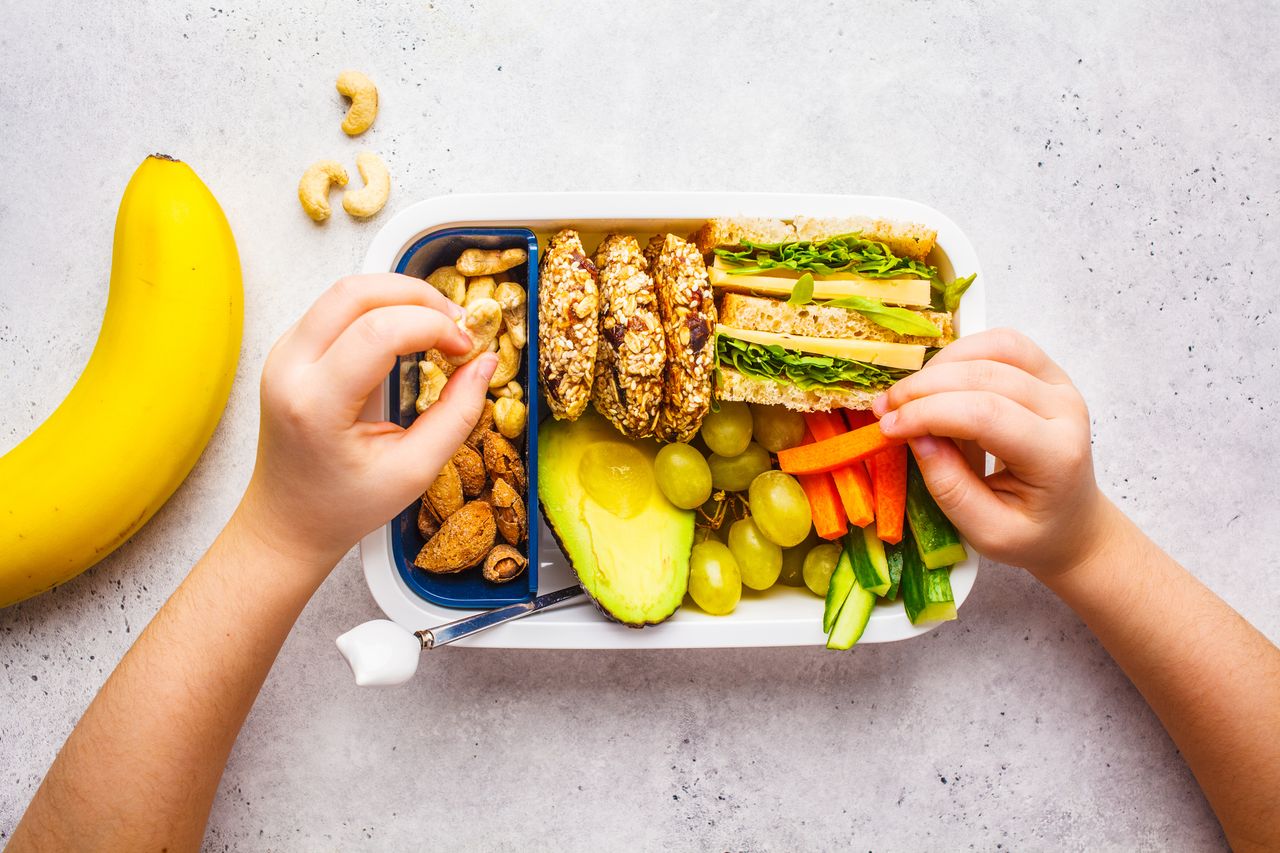 School healthy lunch box with sandwich, cookies, nuts, fruits and avocado on a white background.