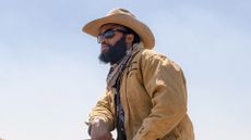 A man sits on a horse in cowboy attire with glasses on 