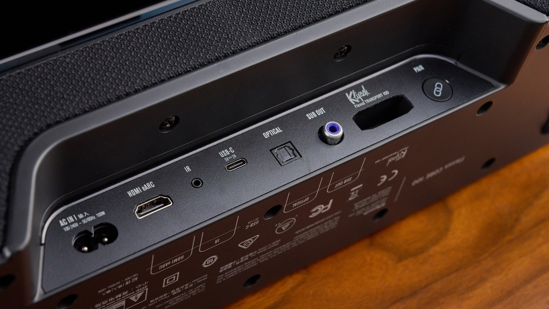 the klipsch flexus core 100 soundbar photographed on a wooden tv stand with a tv behind, showing the remote, buttons, and inputs