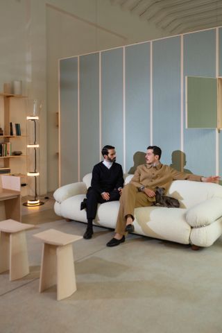 View of Andrea Trimarchi, Simone Farresin and their dog at the Formafantasma Milan studio featuring grey floors, white walls, a white sofa, wooden stools, a wooden bookshelf, a clear tubular floor lamp and a tall blue storage unit