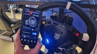 A phone being held next to a Nacon Revosim RS Pure racing wheel with an app on display showing adjustable settings.