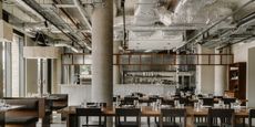 A brutalist restaurant with exposed cement walls, silver ventilation pipes, wooden furniture, and chrome-clad decor is ready for lunch service and brought to life by light.