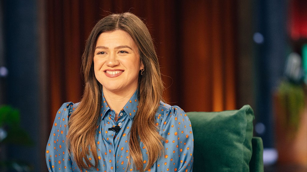 Kelly Clarkson smiles in a blue blouse on the set of The Kelly Clarkson Show.