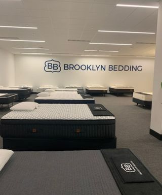 A mattress showroom; mattresses on gray garment against a white wall with the words 'Brooklyn Bedding' written in blue.