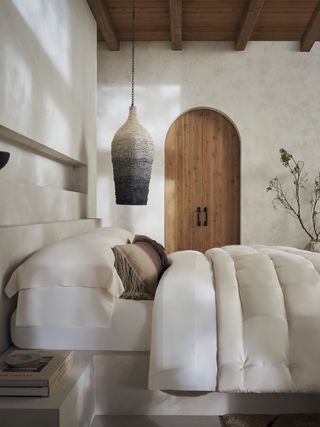 A neutral bedroom with an arched wooden door, white bed spread, and a woven pendant lamp