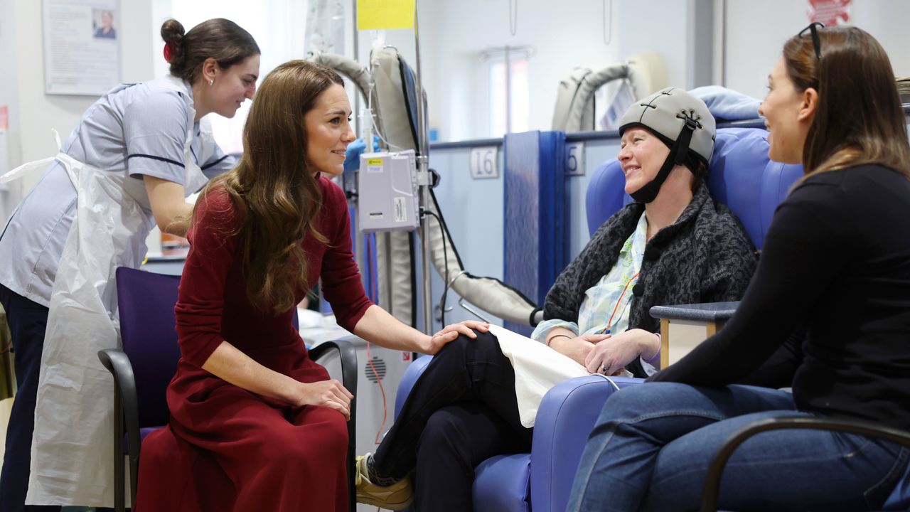 Kate Middleton sitting in a chair wearing a red sweater and skirt putting her hand on the knee of a cancer patient wearing a cold cap getting chemo treatment