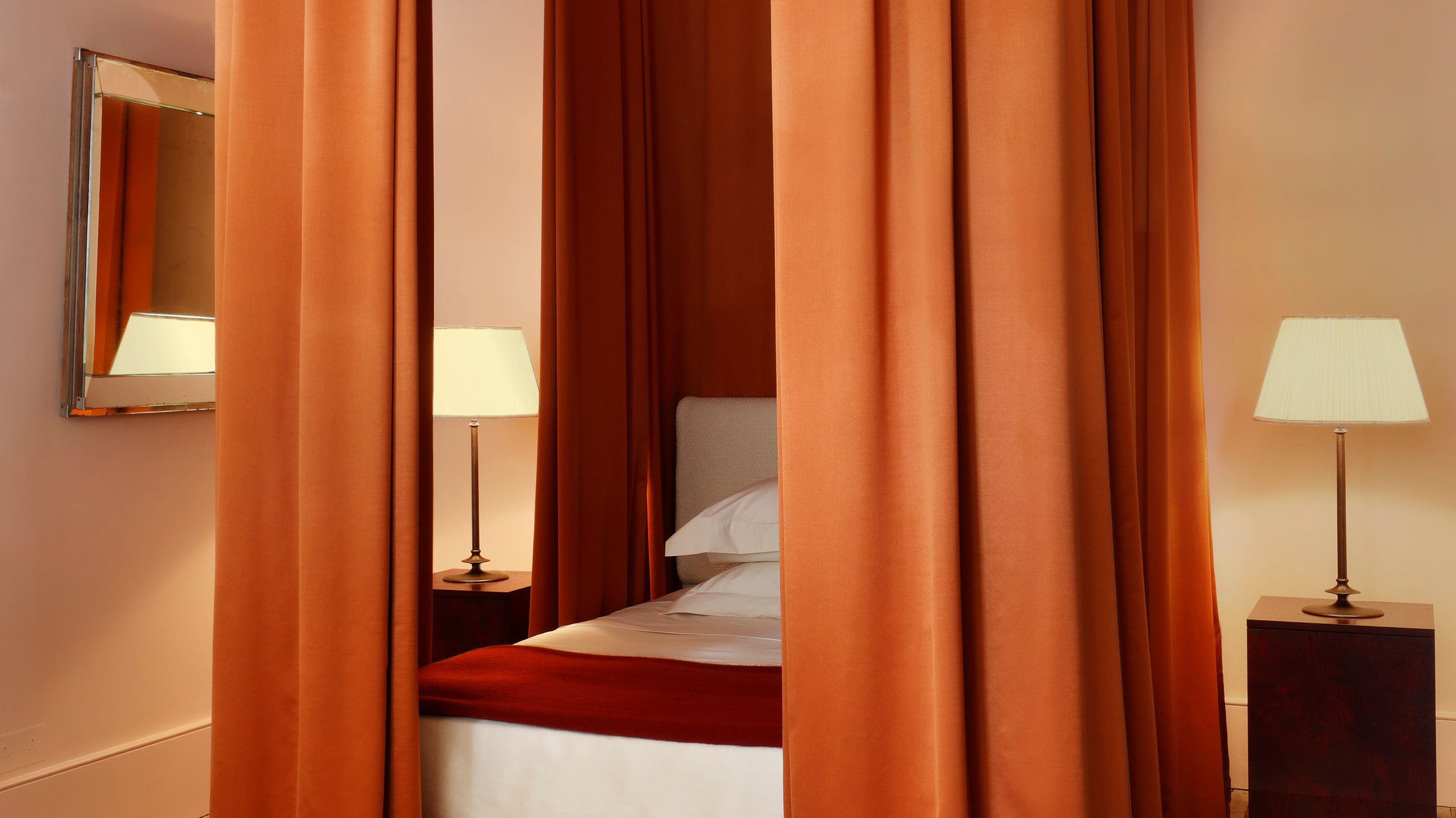 modern bedroom with four poster bed with orange curtains, a mirror on the wall, and wooden plinth side tables with lamps on them