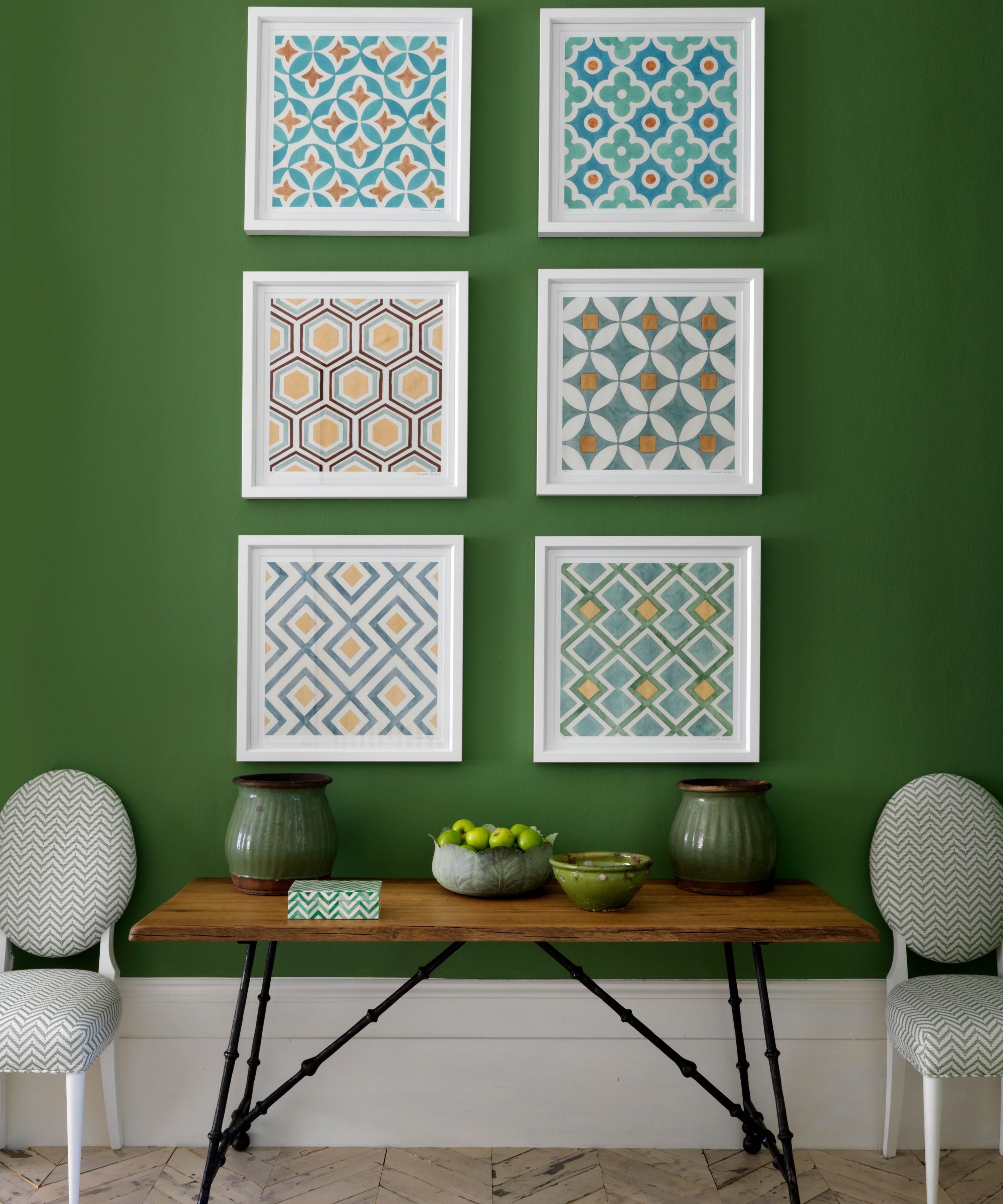 A green wall with framed abstract artwork, a wooden table with green vases and a fruitbowl, and white and green upholstered chairs.