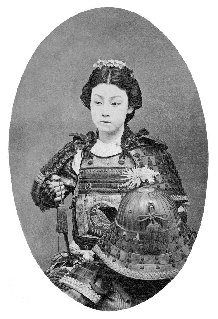 A black and white photograph of a woman wearing traditional samurai armor, sitting and holding a helmet.