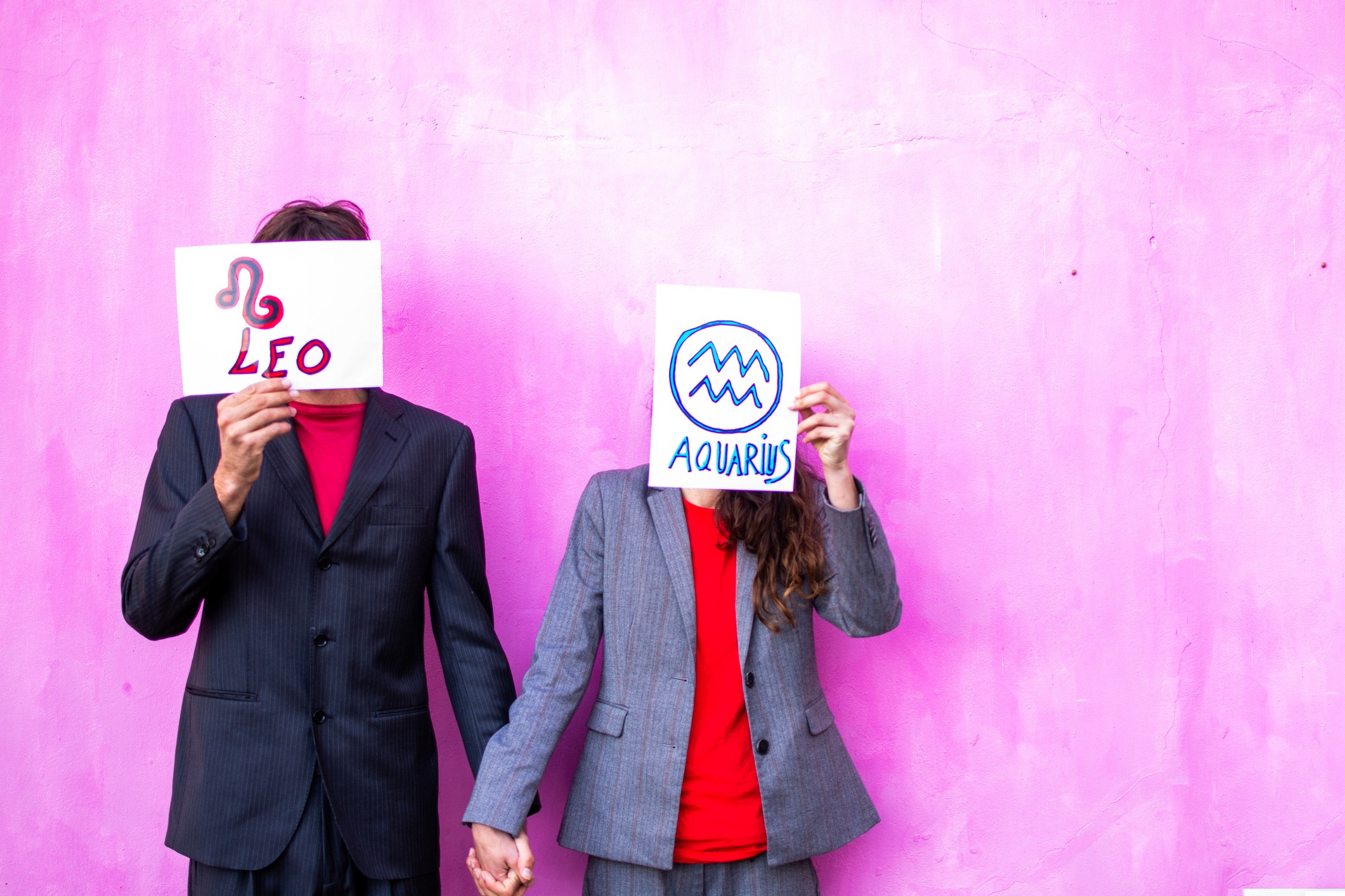 a man and woman holding up bits of paper in front of their faces with their star signs, with a pink background