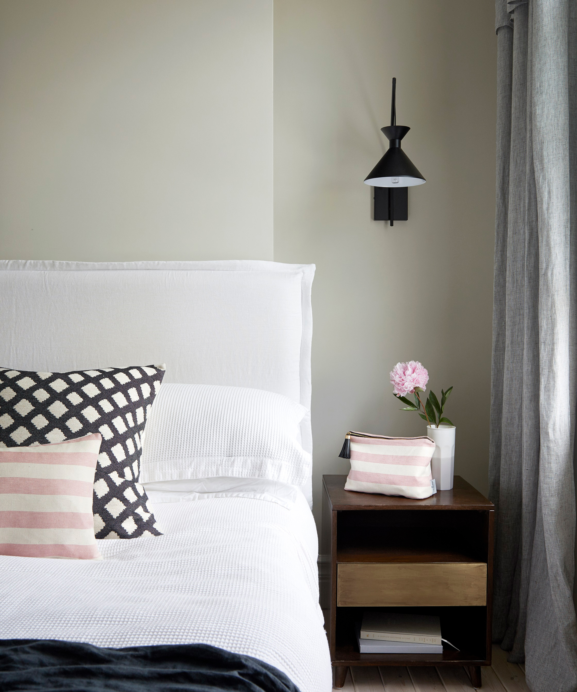 Black and pink bedroom scheme with white padded headboard, light sage walls, peony on bedside, and black wall light.