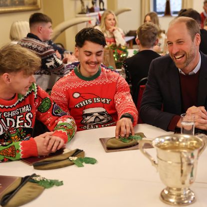 Prince William sitting at a table next to two men in red Christmas sweaters and laughing