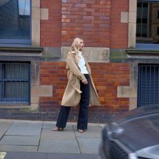 Model wearing a beige trenchcoat, white top, wide leg trousers and heels sold at Debenhams standing on a pavement