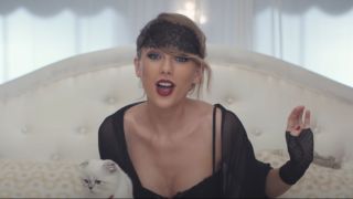 Taylor Swift in a night gown with a black eye mask on top of her head. She's holding her cat while singing Blank Space.