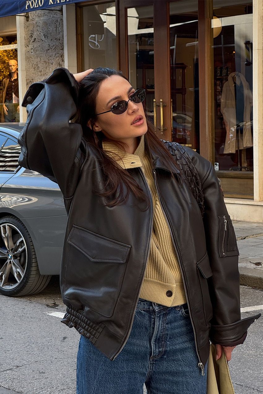 A woman standing outside in the middle of the street wearing a dark brown bomber jacket over a light tan cardigan that's styled as a shirt and paired with dark wash jeans, black wireframe sunglasses, and a tan suede bag.
