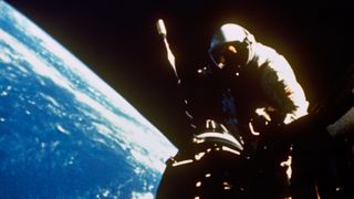 an astronaut floats near a shadowed spacecraft hatch against the black of space. Part of Earth is seen below.