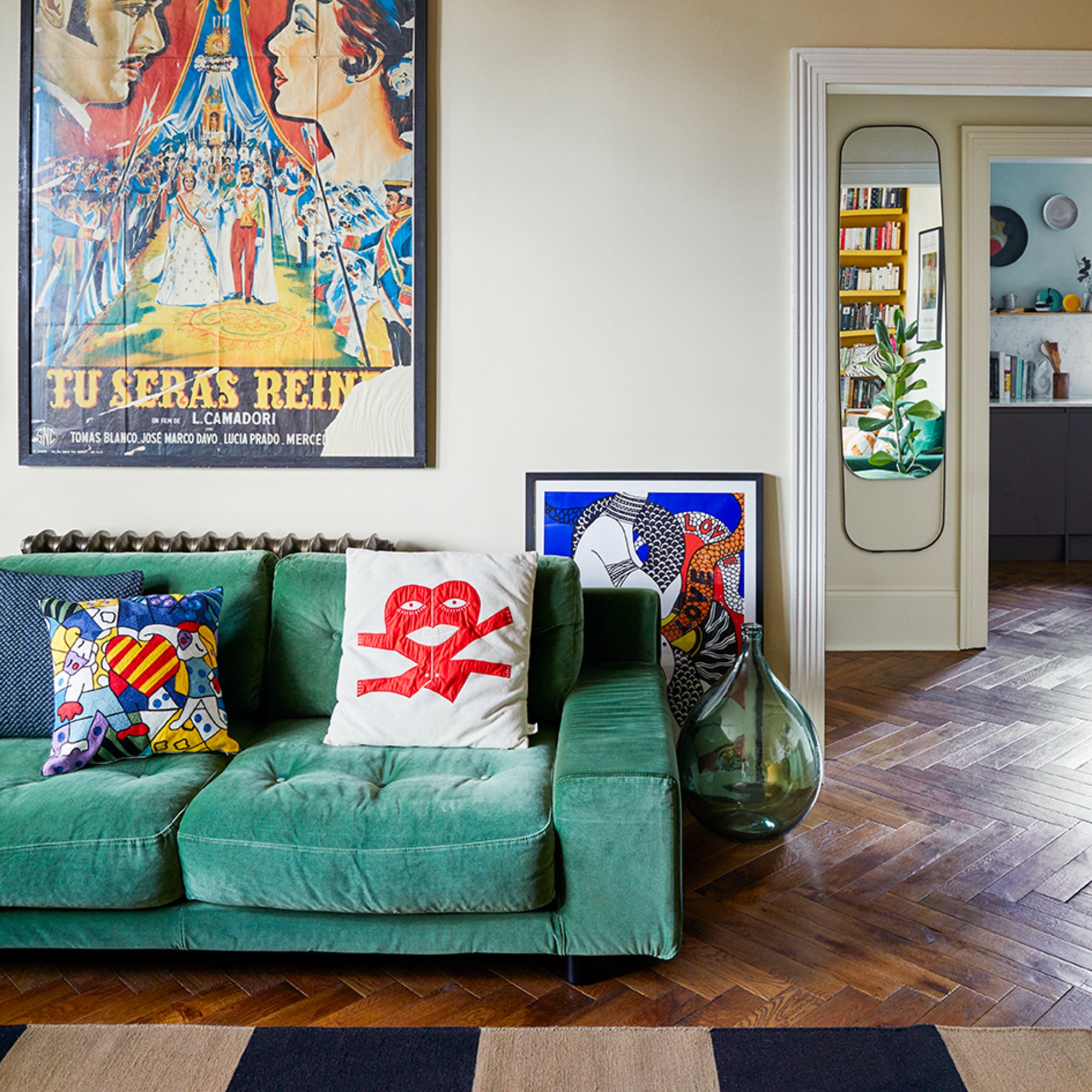 A living room with a green velvet sofa, a chequered rug on a wooden floor, fun cushions and a poster hanging above the sofa