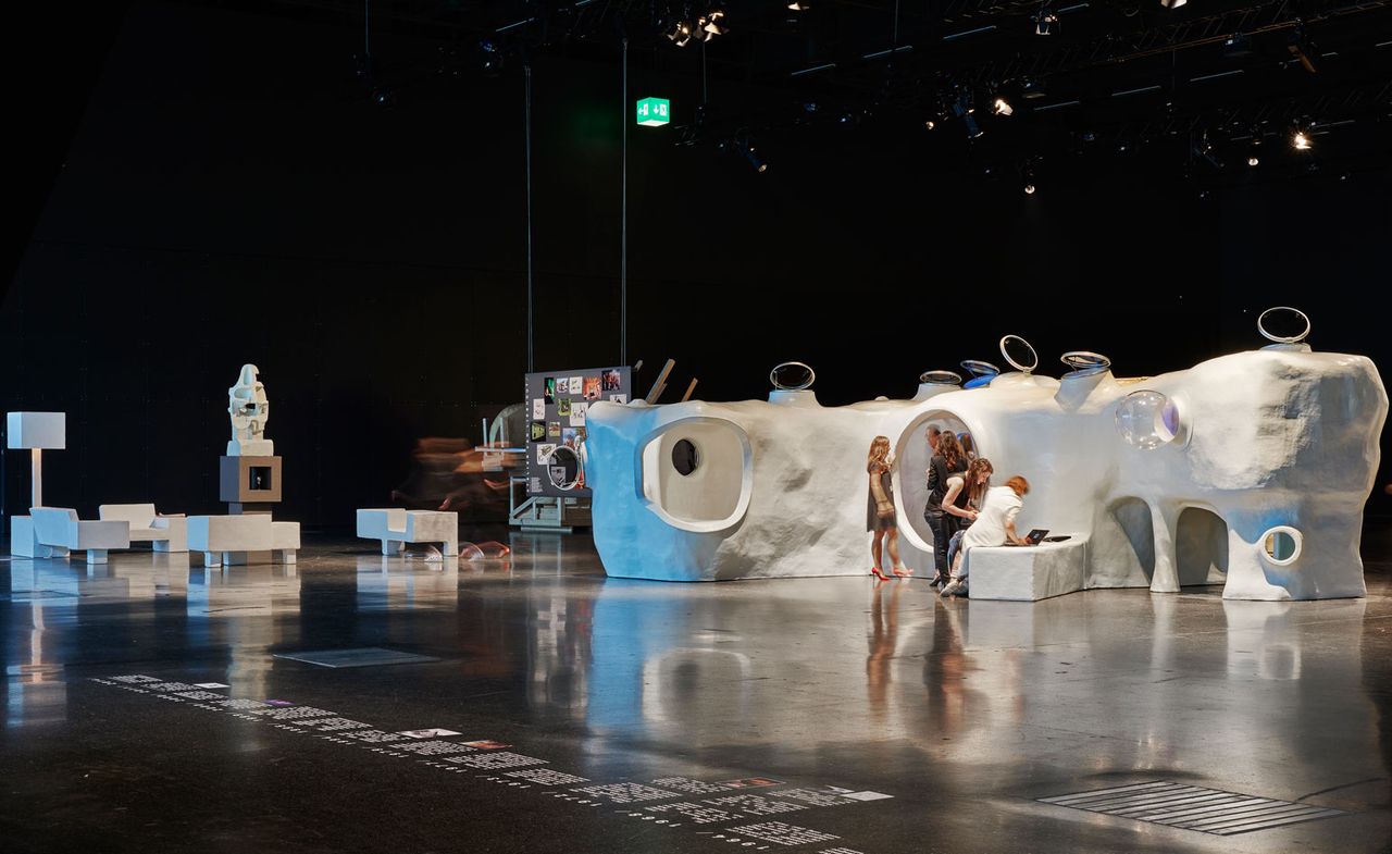 View of the &#039;Design at Large&#039; exhibition featuring a white cave-like structure with a number of people gathered outside it. There is also a grey board with images suspended from the ceiling and white seating along with other pieces displayed in a space with a black floor and background