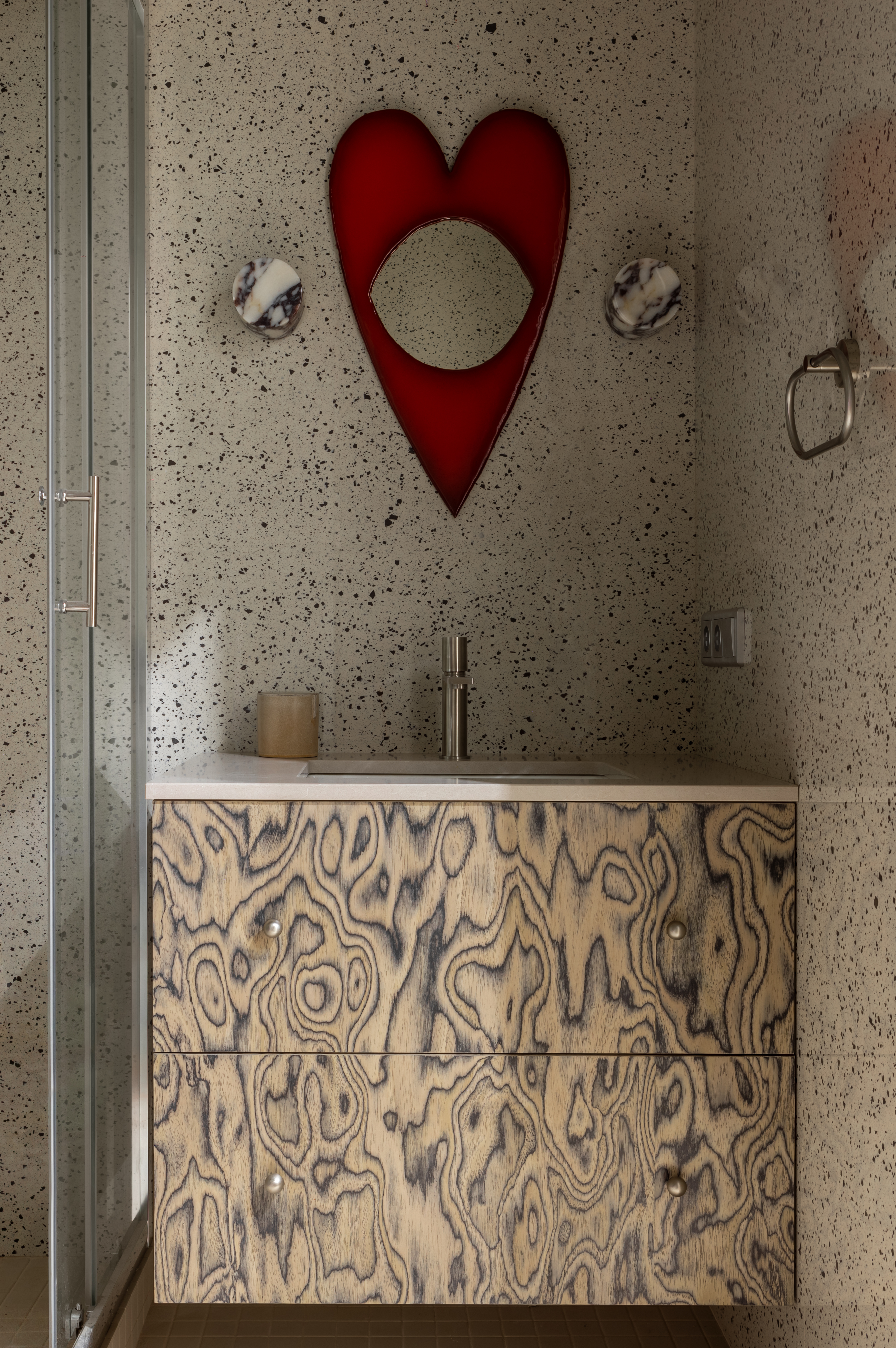 A bathroom with terrazzo walls, wood veneer vanity, and heart-shaped mirror 