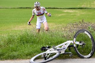 Tadej Pogacar gets up amid tall grass and brambles and heads back toward his bike lying on the side of the road after a crash