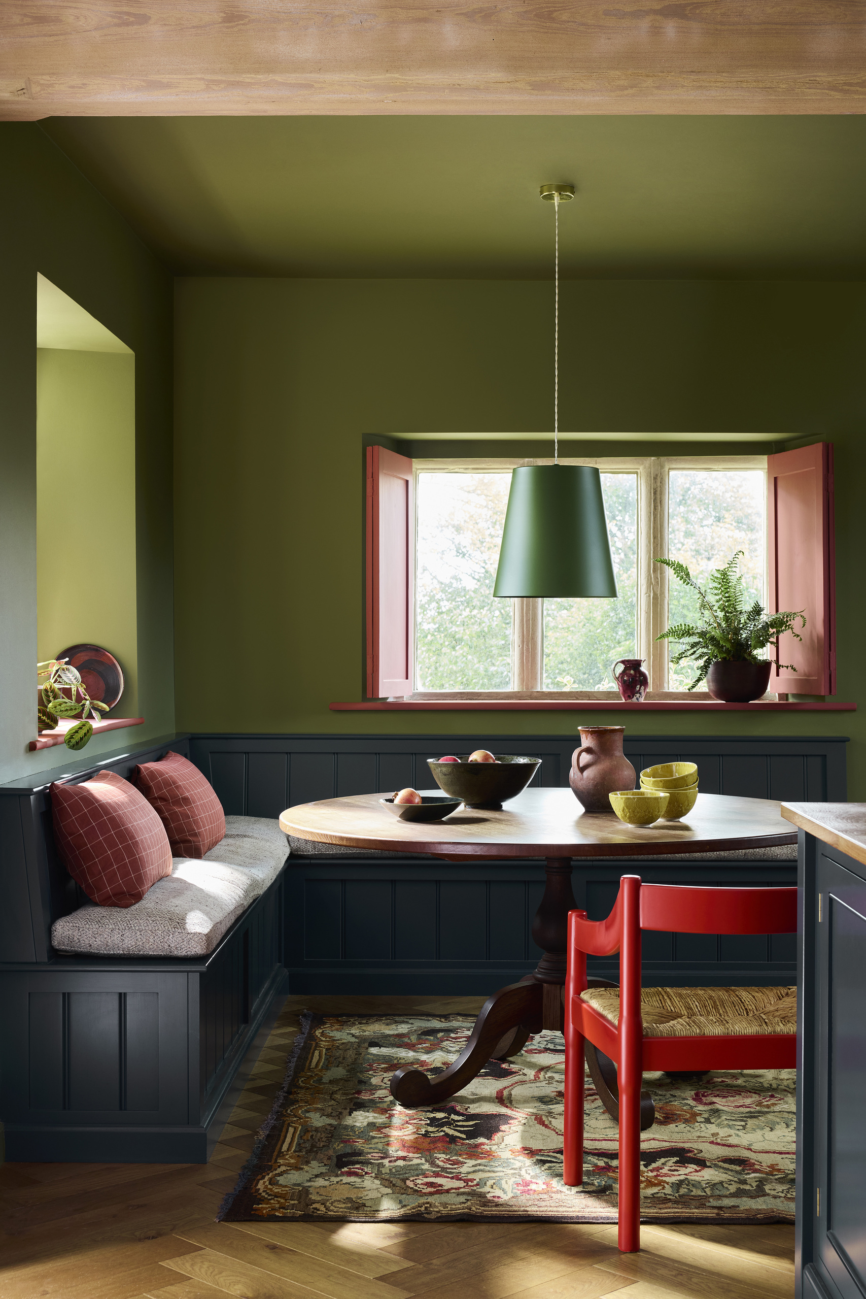Image of a dining nook with olive green walls and a navy paneled bench seat. There is a round table, a red chair, and red trim on the window