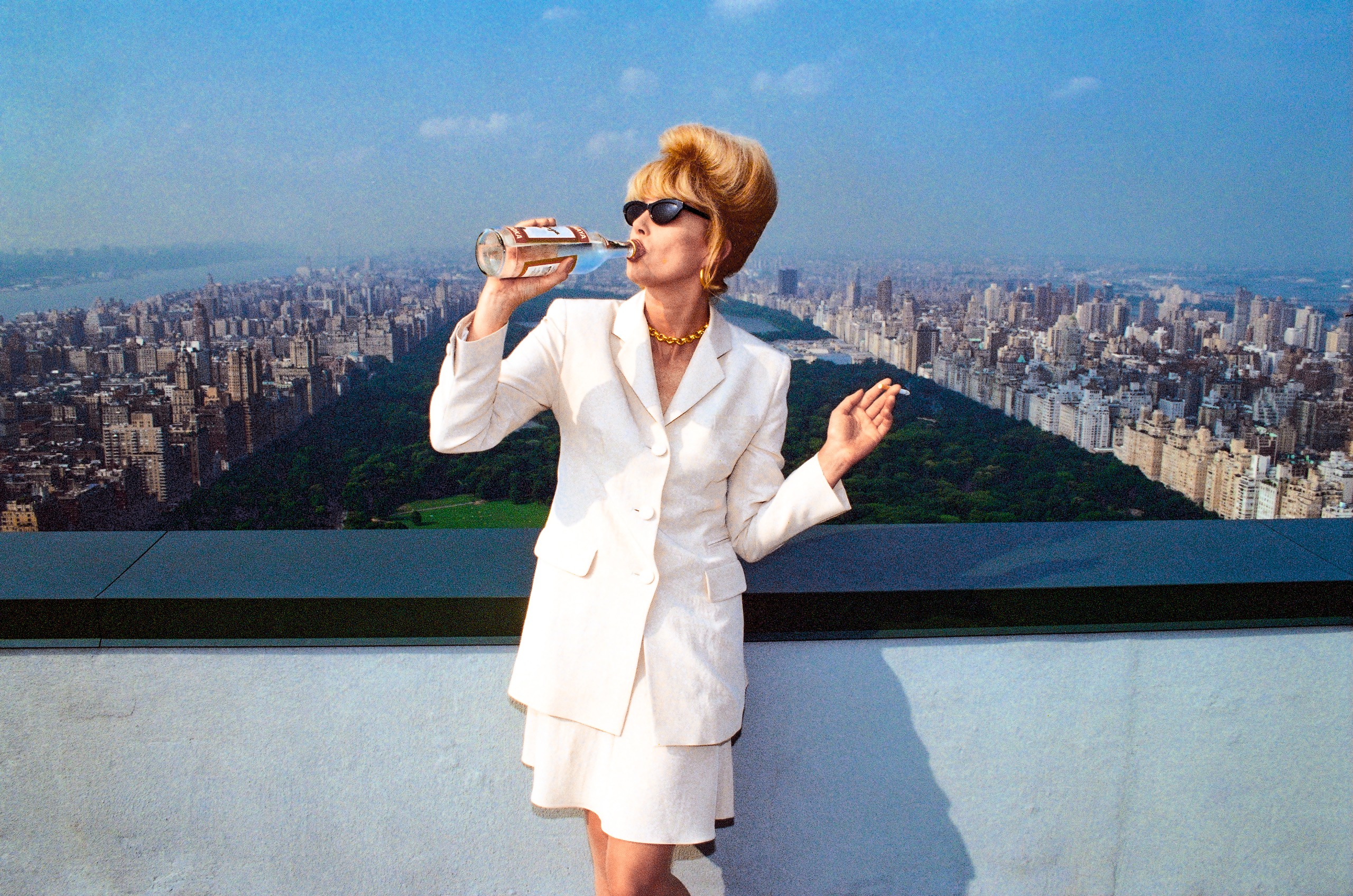 Color photo of Joanna Lumley in a white skirt suit, sunglasses and a blonde beehive, swigging from a vodka bottle with a cigarette in her other hand on a rooftop overlooking Central Park
