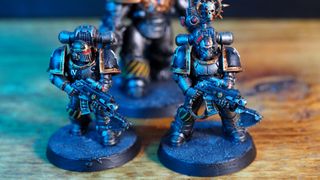 Two MKII Crusader Pattern Space Marines on a wooden surface, with a Saturnine Terminator behind them