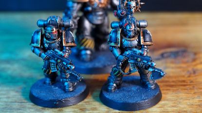 Two MKII Crusader Pattern Space Marines on a wooden surface, with a Saturnine Terminator behind them