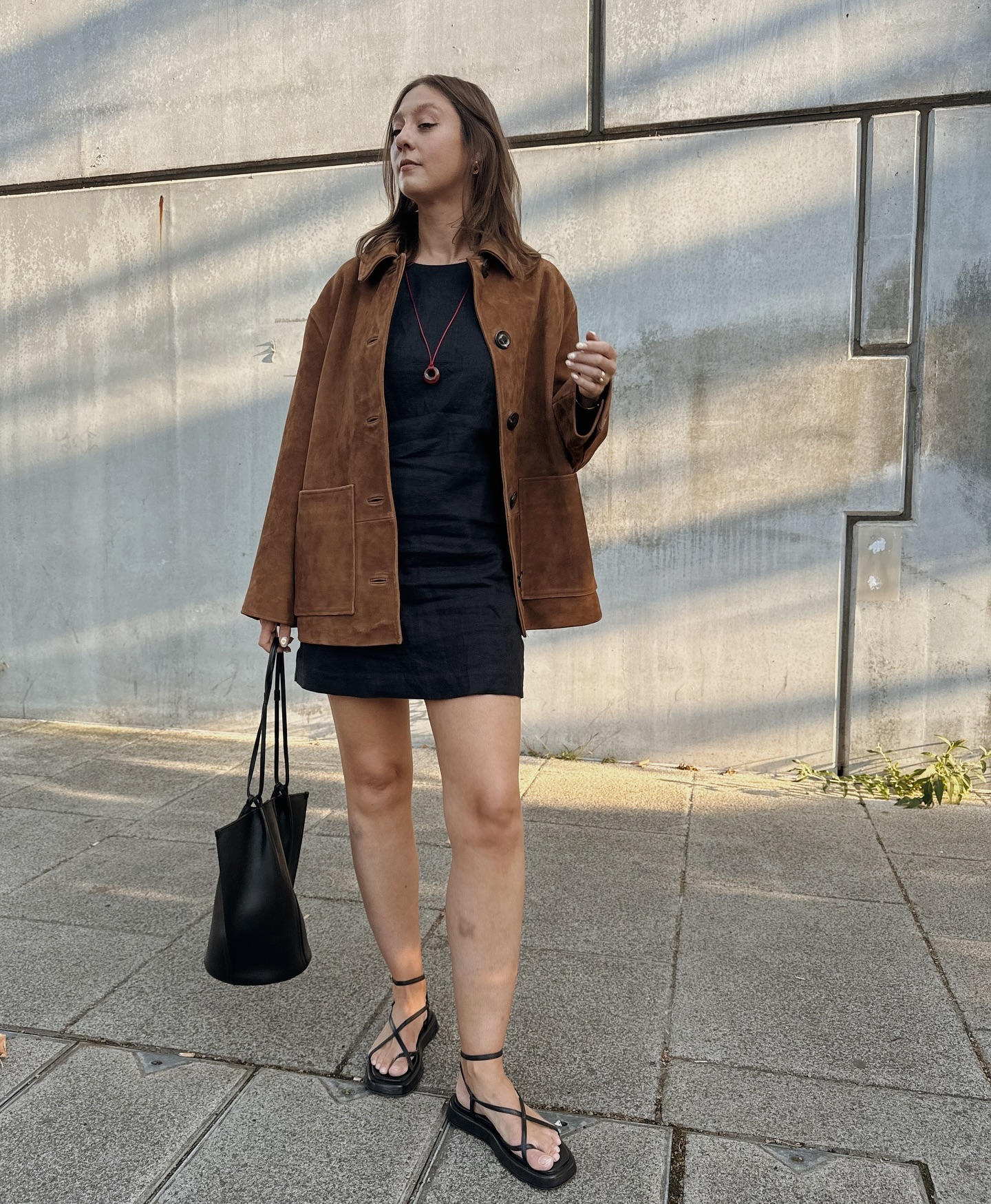 Woman wears a brown suede jacket, black mini dress, black sandals and carries black tote bag