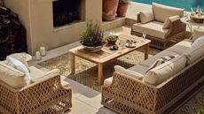 Wicker patio furniture surrounding a wooden coffee tale, in front of a stone outdoor fireplace, beside a pool.