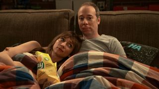 Lauren Lapkus and Kevin Susssman snuggle on a couch with a plaid blanket. There's an open bag of chips in between them.