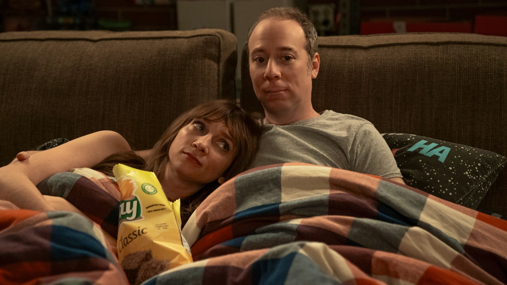 Lauren Lapkus and Kevin Susssman snuggle on a couch with a plaid blanket. There's an open bag of chips in between them.