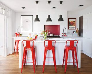 A white kitchen with red bar stools around the island with trio of black pendant ceiling lights