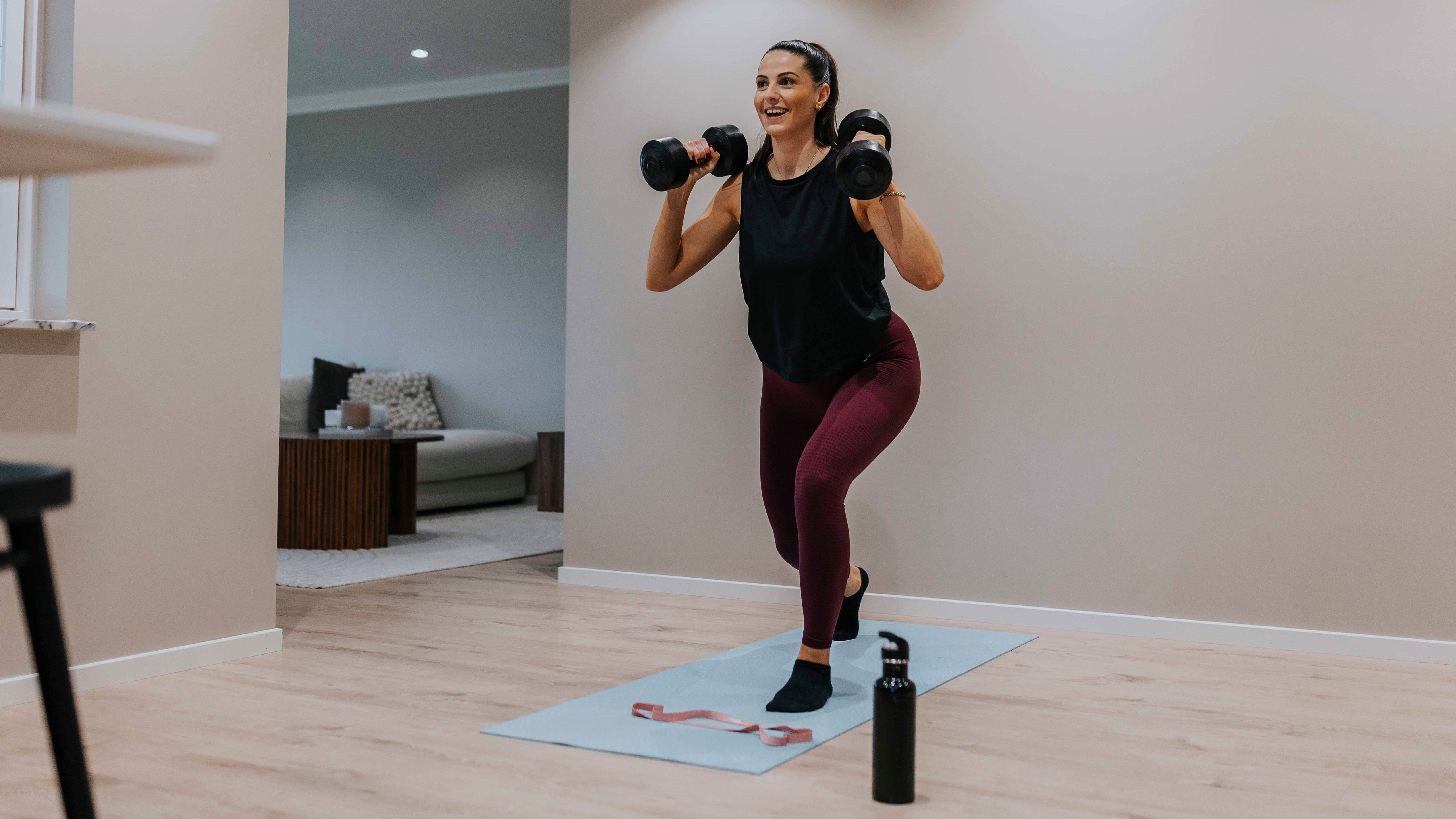 A woman stands holding dumbbells at her shoulders. She is stepping back with her right leg and exercising on a mat with a water bottle nearby.