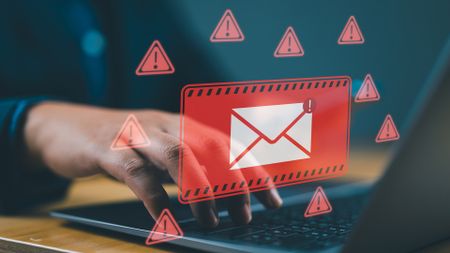 An email symbol inside a red square warning sign, surrounded by red triangles with exclamation marks inside them, superimposed on someone typing on a laptop