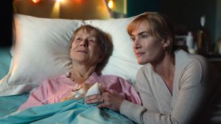 Helen Mirren laying down in a hospital bed smiling (but looking sick) as June and Kate Winslet as her daughter Julia next to her smiling in Goodbye June 
