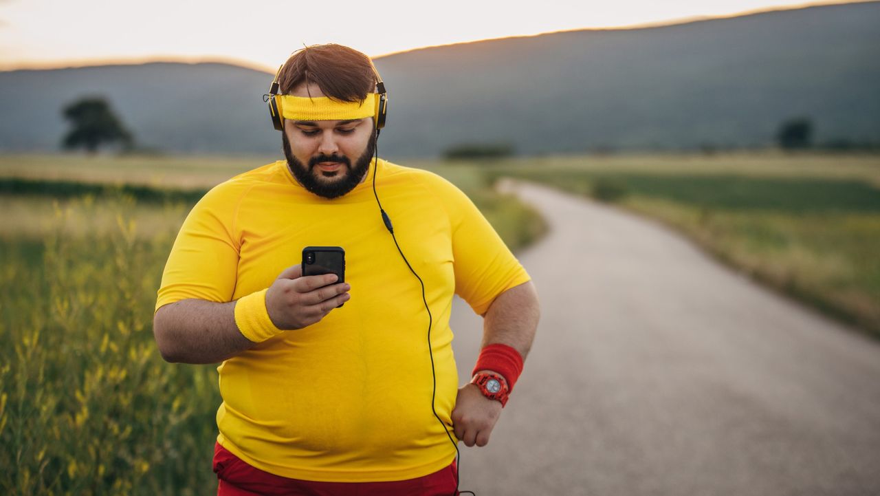 5 easy strategies to get fit if you&#039;re lazy: Pictured here, smiling overweight man wearing sports gear looking at his smartphone on the road