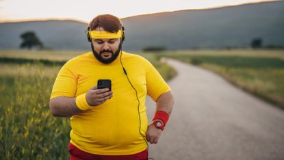 5 easy strategies to get fit if you're lazy: Pictured here, smiling overweight man wearing sports gear looking at his smartphone on the road