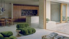 A spacious living room with green plush couches and a coffee table with books, an open concept kitchen and dining room opening out into an outdoor space