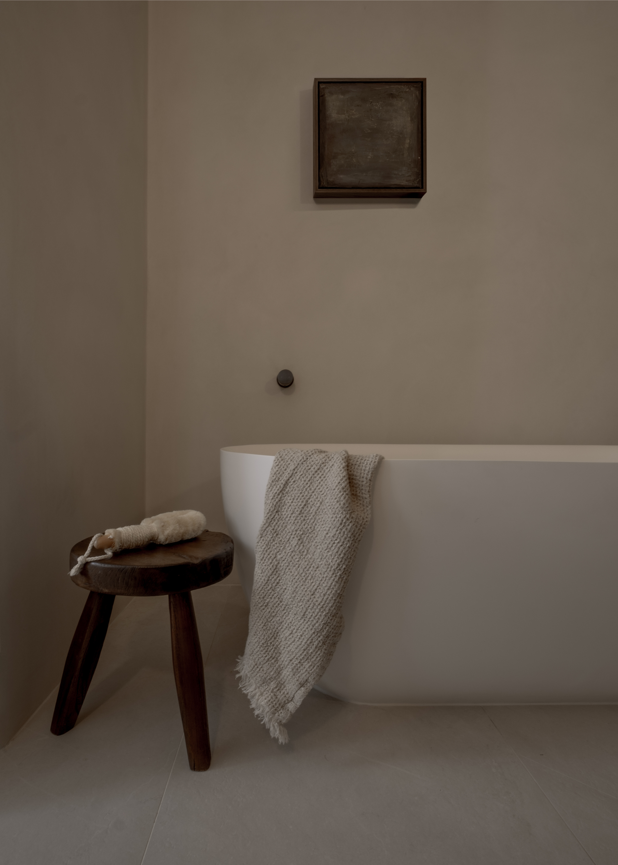 An off-white bathroom with a standing white bath tub, a textured towel, a wood stool, and a scrubber