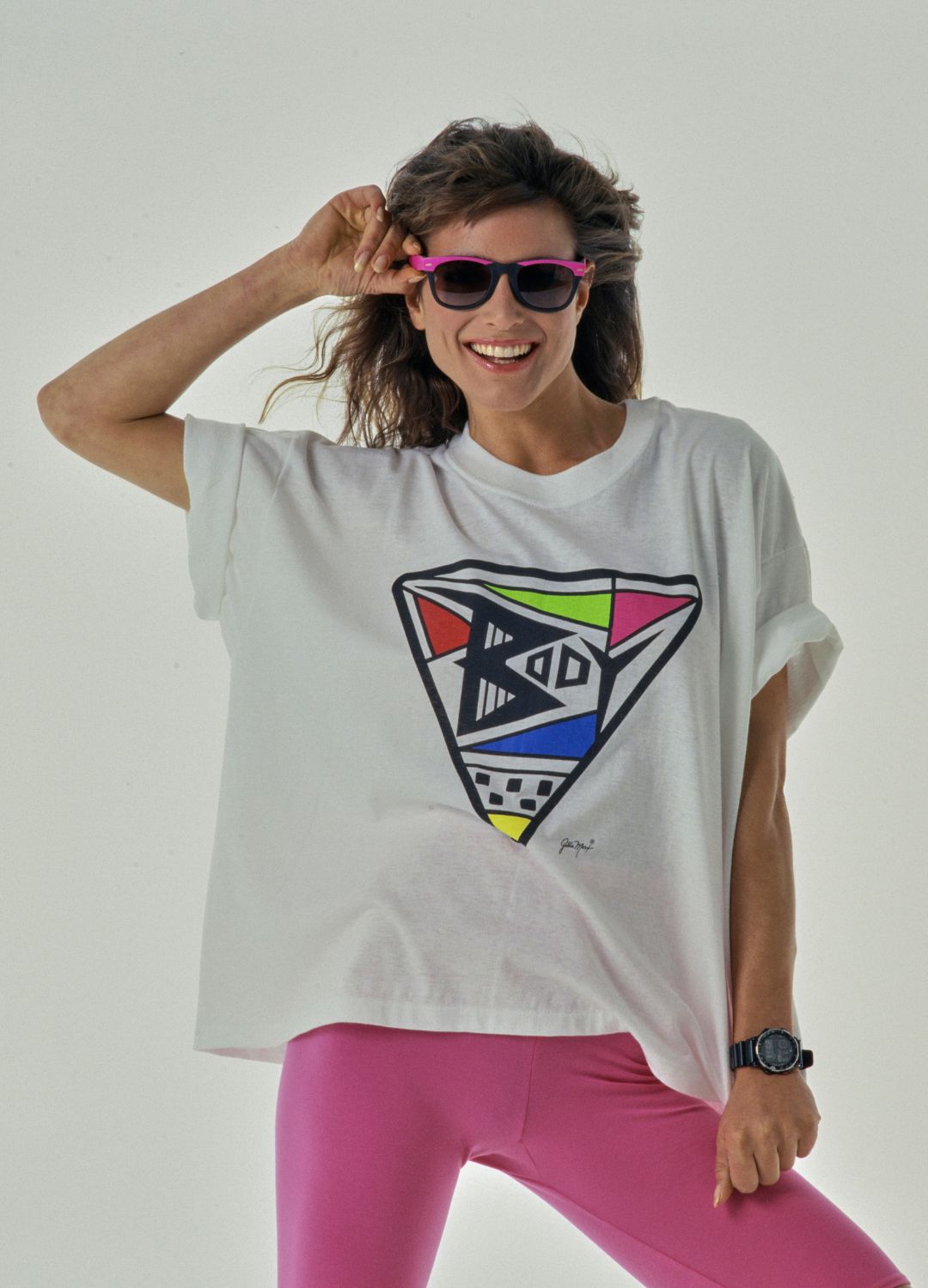 A woman modelling a white t-shirt featuring an abstract design round the word 'Body', and pink cycling shorts, her right hand holding the frame of her sunglasses in a Summer studio fashion shoot, United States, 1989.