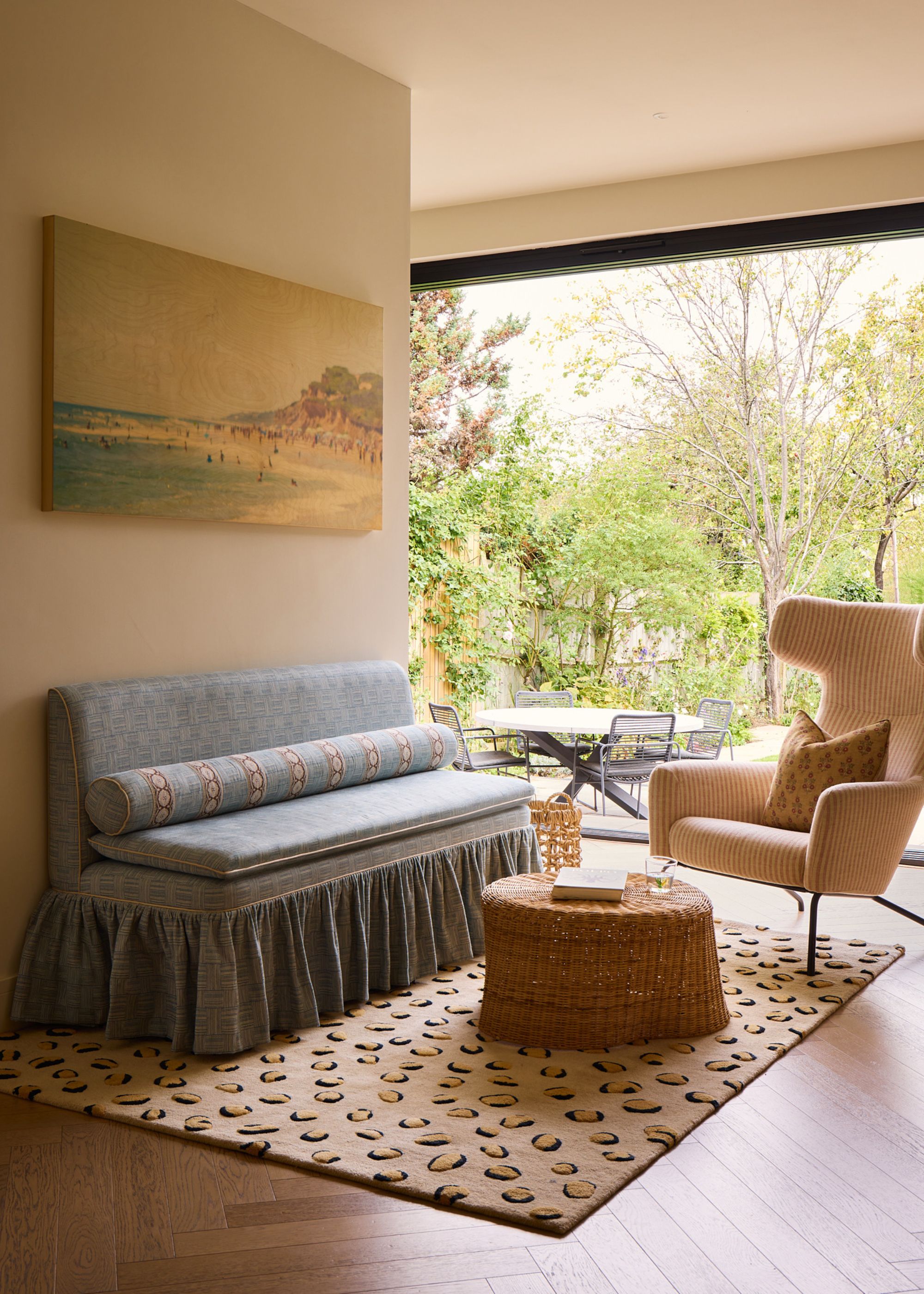 A modern living space in a new home with a large glass wall with views to the garden, light beige walls with a landscape artwork, a small blue sofa with a patterned rug, a woven coffee table, and a neutral armchair.