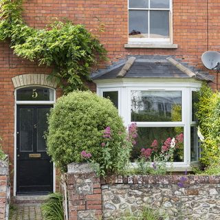 house exterior with red brick wall and plants tress and white window with black door with white frame