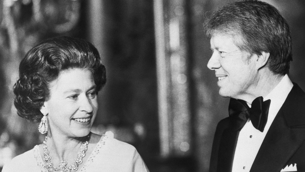 A black and white picture of Queen Elizabeth wearing diamond jewels and a white dress and Jimmy Carter wearing a tux at Buckingham Palace