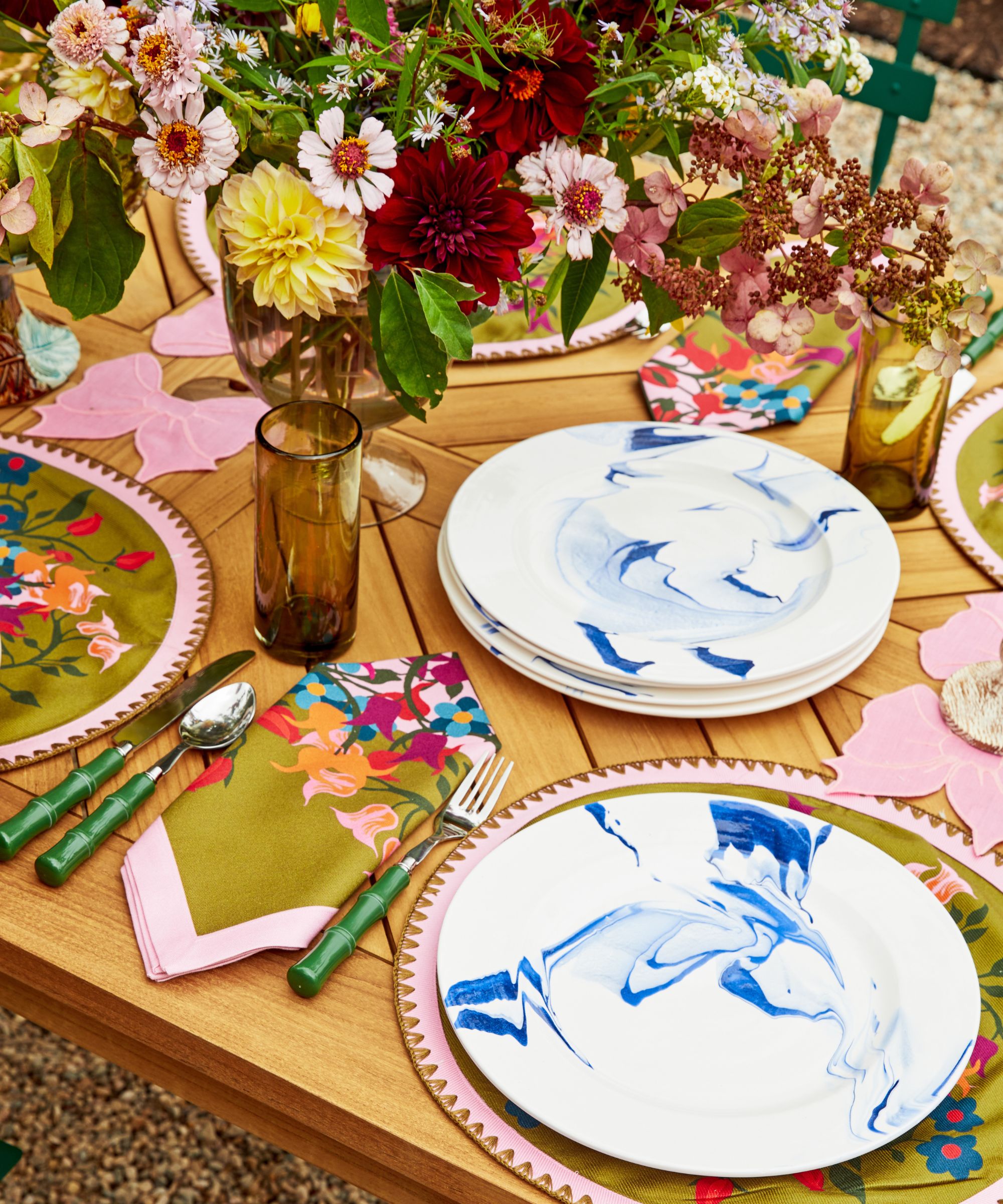 Wooden table with colourful placemats, green bamboo cutlery, white and blue plates, floral napkins, and amber glasses with a large floral arrangement at the centre.