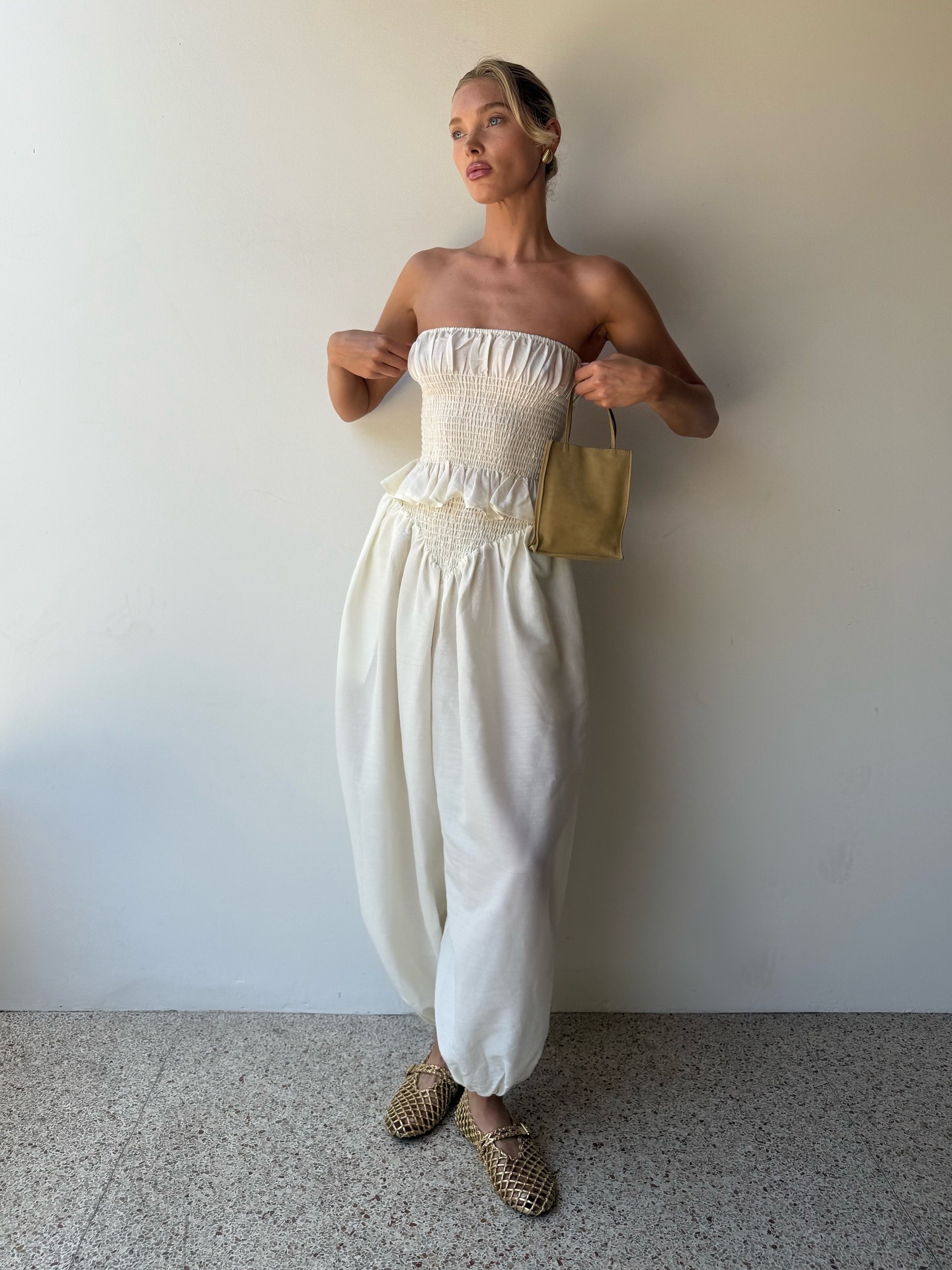 Elsa Hosk stands inside against a white wall wearing a white strapless two-piece with gold woven flats from Alaia.
