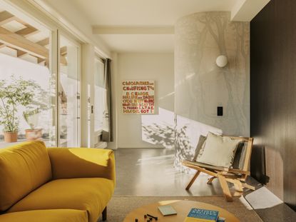 A bright lounge area at the hotel with a mustard yellow couch situated next to the floor-to-ceiling windows, across from which is a coffee table and a wooden chair with pillows.