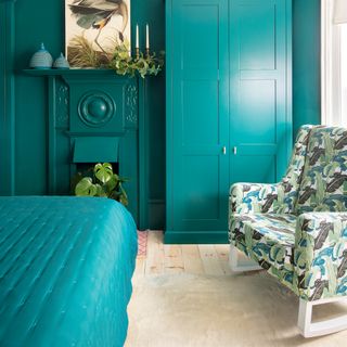 Bold teal bedroom with painted wardrobes and fireplace and a botanical pattern armchair