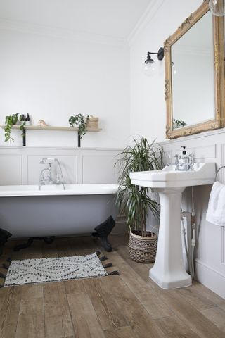 Bathroom with light grey panelling and white walls, wood floor, grey roll top bath, white Edwardian-style sink and gold ornate mirror