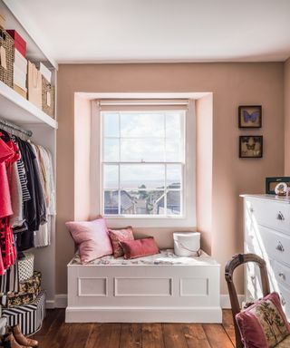 A pastel pink closet area with white window bench seat, white chest of drawers and an open clothes rail.